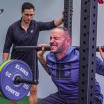 Muscular sportsman in activewear squatting with heavy barbell with effort while listening trainer in gym