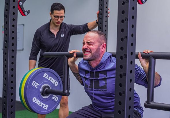 Muscular sportsman in activewear squatting with heavy barbell with effort while listening trainer in gym