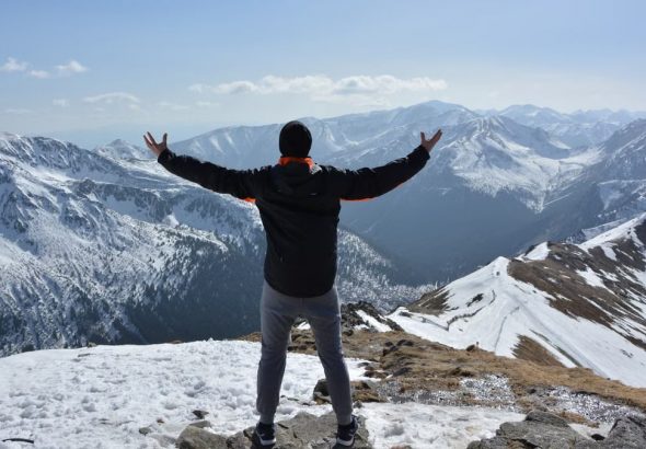 A man stands on a snowy mountain peak with arms raised, embracing the breathtaking view.